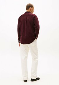 Bordeaux corduroy shirt met een button-down kraag en lange mouwen, gedragen met witte broek en zwarte schoenen, gezien van achteren.