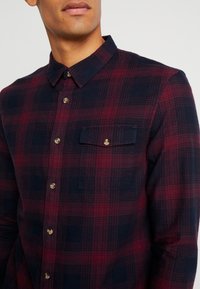 Uomo che indossa una camicia a quadri rossa scura e navy con bottoni, dotata di una tasca sul petto e colletto, mostrato dalla nuca a metà torace.