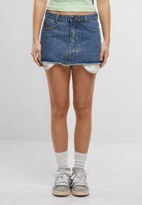 Denim mini skirt with a frayed hem, light blue color, five-pocket design, and a front button closure. Worn over white shorts and paired with gray sneakers.