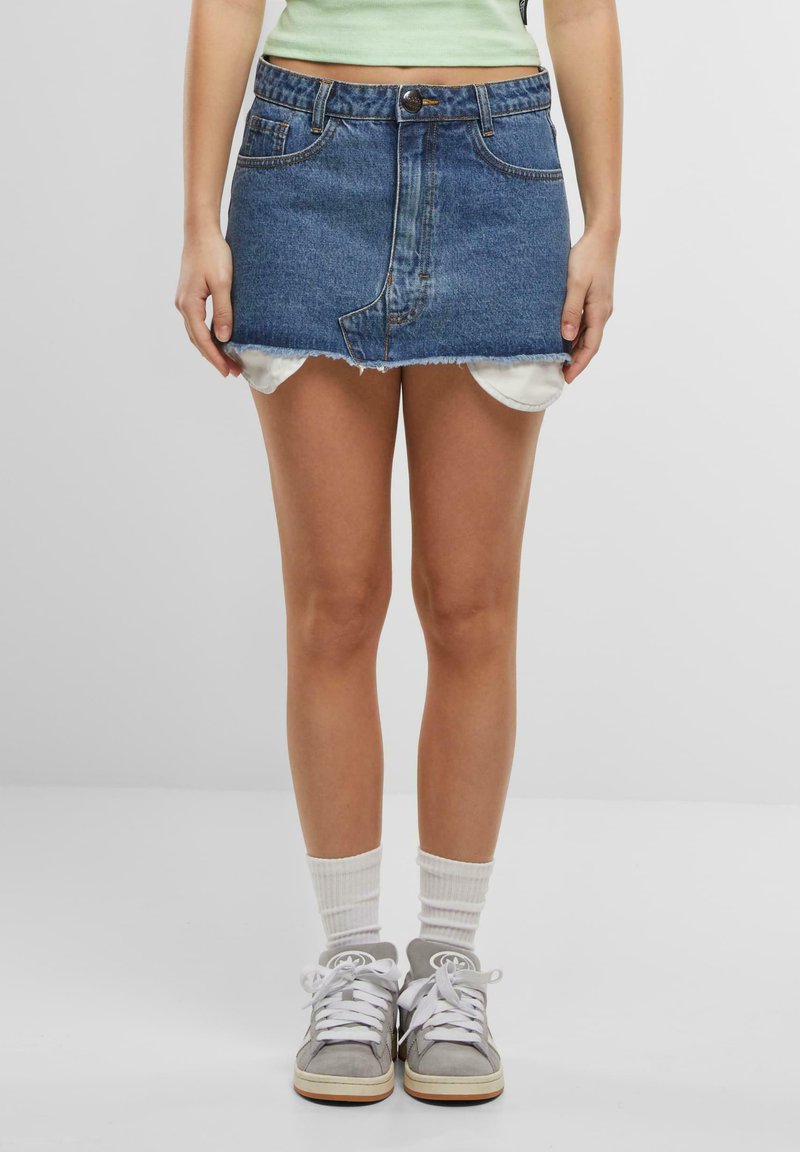 Denim mini skirt with a frayed hem, light blue color, five-pocket design, and a front button closure. Worn over white shorts and paired with gray sneakers.