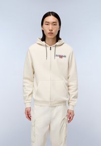 Felpa con zip color crema, con tasca frontale, cappucci grigi e un logo con accentuazioni rosse e blu. Indossata con pantaloni chiari abbinati.