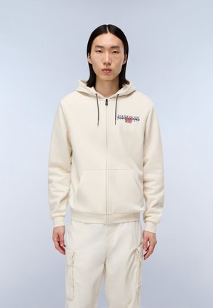Sudadera cremosa con cremallera y bolsillo frontal, cordones grises y un logo con acentos rojos y azules. Combinada con pantalones ligeros a juego.