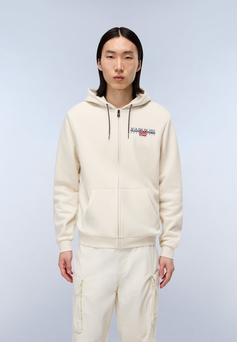 Felpa con zip color crema, con tasca frontale, cappucci grigi e un logo con accentuazioni rosse e blu. Indossata con pantaloni chiari abbinati.