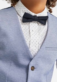 Veste bleu clair texturée avec un nœud papillon bleu foncé sur une chemise blanche ornée d'un motif triangulaire noir subtil et de détails de boutons.