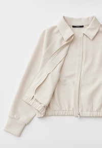 Giacca beige con zip, collo, vita elasticizzata e maniche lunghe, parzialmente aperta su sfondo bianco.