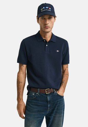 Donkerblauwe polo met korte mouwen en een kraag. Bevat een klein geborduurd logo op de borst. Gemaakt in combinatie met donkerblauwe jeans en een bruine riem.