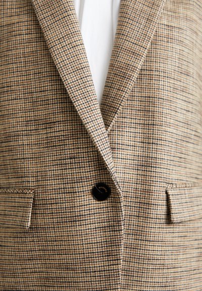 Brun og beige ternet blazer med en sort knap og to frontlommer med klap, båret over en hvid skjorte.
