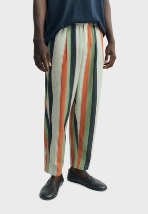 Muškarac obučen u široke hlače s uspravnim prugama u zelenoj, narančastoj, bež i tamno plavoj boji, uparene s crnim obućama bez vezica.