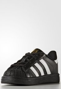 Svarta sneakers med en texturerad gummitå, tre vita ränder och snörningsdesign. Har en svart mesh-häl och en guldfärgad logotyp på tungan.