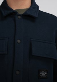 Marineblauwe button-up jas van zacht materiaal met twee borstzakken, zwarte knopen en een logo patch op de linker zak, voorzien van een opgestikte kraag.