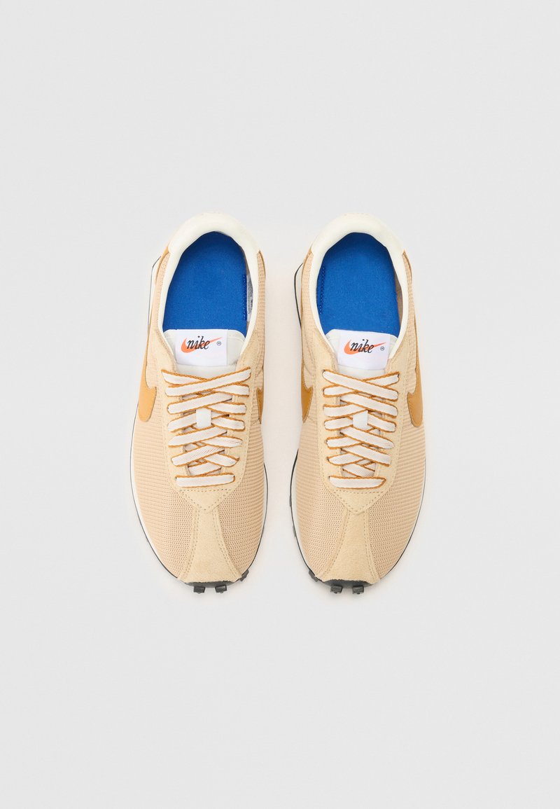 Baskets de sport beiges avec une tige en maille, semelle intérieure bleue, logo Nike blanc et accents orange. Présentent des lacets ronds et une semelle texturée.
