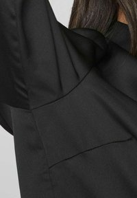 Schwarze Bluse aus Stoff mit glatter Textur, die über strukturierte Schultern und Nahtdetails verfügt. Keine sichtbaren Muster oder Beschläge.