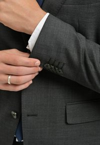 Grå kavajsärm med fyra svarta knappar och dekorsöm. Vit skjortärm synlig nedanför. Slätt tyg med fin textur.