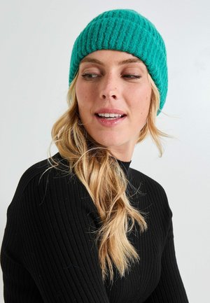 Gorro de punto en color verde azulado con textura acanalada, diseño ajustado y color sólido. Llevado con una blusa negra de manga larga.