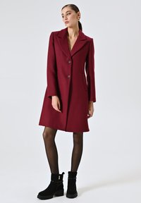 Bordeaux wollen blend jas met een geknokte kraag, twee knopen, zijzakken en een knie-lange A-lijn silhouet. Gecombineerd met zwarte enkelboots.