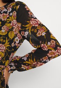 Chemise en tissu noir transparent ornée de motifs floraux en rose et jaune, avec manches longues et poignets froncés, mettant en valeur des dessins botaniques détaillés.