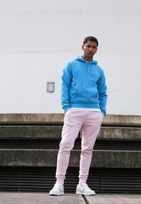 Lichtblauwe hoodie met een kangoeroezak, gecombineerd met roze sweatpants. Het model draagt witte sneakers met blauwe en gele accenten, terwijl ze op de trappen staat.