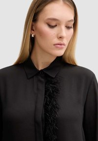 Blusa nera con colletto e dettaglio verticale piumato sulla parte anteriore. Tessuto liscio, chiusura a bottoni e vestibilità sartoriale.