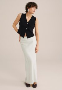 Crna bezrukavna prsluk s V-izrezom i srebrnim gumbima, uparen s kremasto dugom suknjom i tamnosmešnim cipelama na slip-on. Glatka tekstura.