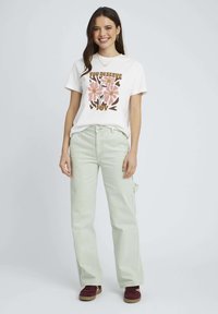 T-shirt blanc avec un motif floral en rose et marron, associé à un pantalon large vert clair. Modèle portant des baskets bordeaux, debout.