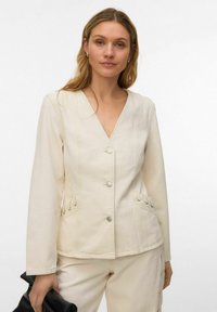 Femme portant une veste crème boutonnée avec un détail à lacets sur les poches et un pantalon assorti, tenant une pochette noire dans sa main gauche.