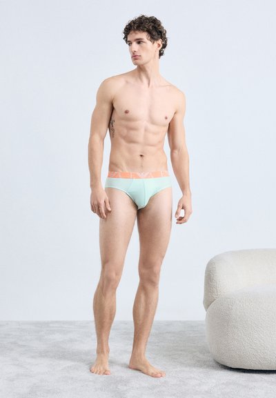 Slip bleu clair avec ceinture orange, arborant un logo. Le modèle se tient sur un tapis texturé, affichant une silhouette ajustée.