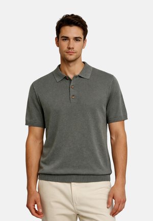 Giovane uomo che indossa una polo a maniche corte grigia a maglia con bottoni, abbinata a pantaloni beige chiaro, in piedi davanti a uno sfondo semplice.