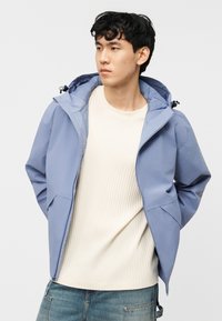 Νεαρός άνδρας με κοντά μαύρα μαλλιά, φοράει ελαφρύ γαλάζιο μπουφάν με κουκούλα πάνω από ένα εκρού πλεκτό πουλόβερ και μπλε τζιν, κοιτάζει προς τα πλάγια.