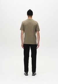Khaki katoenen T-shirt met een klassieke pasvorm, gecombineerd met zwarte broeken, gezien vanuit de rug. Een zwarte beanie wordt op het hoofd gedragen.