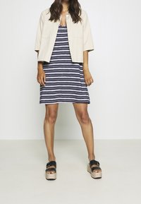 Veste beige clair à manches courtes portée sur une robe rayée bleu marine et blanc. Le modèle porte des sandales plateformes noires avec une semelle en liège.