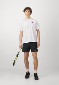 T-shirt bianca in cotone con un piccolo logo colorato, pantaloni sportivi neri e sneakers nere, mentre tiene una racchetta da tennis con un'impugnatura bianca.