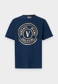 Navy zila kokvilnas T-kreklu ar apaļu kakla izgriezumu un īsām piedurknēm. Tam ir liels zelta apļveida logotips ar tekstu "VERSACE JEANS COUTURE".