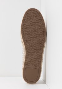Semelle d'une chaussure beige avec une semelle en caoutchouc brun texturé, ornée de motifs répétitifs de logos "MK", présentée sur un fond blanc dans un coin.