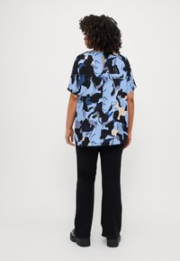 Camicia a maniche corte con un motivo astratto blu, nero e beige, dotata di un dettaglio a chiave sul retro, abbinata a pantaloni neri svasati.
