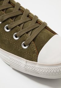 Sneaker verde in suede con punta e suola in gomma bianca, dotato di occhielli metallici e lacci piatti. Superficie testurizzata con dettagli di cucitura marrone.
