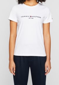 Kobieta ubrana w białą koszulkę z krótkim rękawem z logo "Tommy Hilfiger Est. 1985" oraz granatowe spodnie na jednolitym tle.