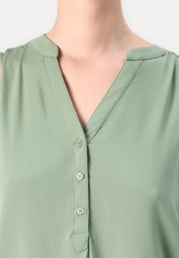 Grønn, ermeløs bluse med V-hals, med en knappestolpe og myk, glatt stoff. Minimalistisk design og subtil tekstur.