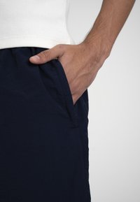 Main masculine reposant dans la poche avant d'un pantalon bleu foncé, portant une chemise blanche côtelée sur un fond uni.