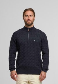 Navyblauwe kabelgebreide trui met een hoge kraag, halve ritsluiting, geribde manchetten en zoom. Heeft een klein logoaccent op de borst.