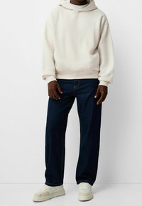 Sudadera beige clara con un bolsillo tipo canguro, combinada con jeans de pata ancha azul oscuro y zapatillas color crema. Textura de tela suave en todo.