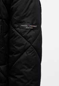 Zwarte gewatteerde pufferjack met een stoffen label met het logo "FRIEDA FREDDIES NEW YORK", weergegeven op de bovenste mouw. Getextureerd oppervlak.
