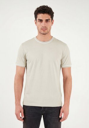 Mand står frontalt, iført en lysebeige t-shirt med korte ærmer og mørke jeans, med neutralt udtryk foran ensfarvet baggrund.