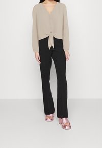 Femme portant un chemisier beige à manches longues boutonné avec un nœud à l'avant, un pantalon noir droit, et des sandales à talons blocs métalliques roses, debout.