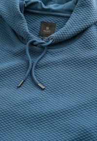 Felpa blu con texture e motivo trapuntato, con cappuccio regolabile tramite cordoncino e estremità in metallo. Etichetta con la scritta "NEXT" è visibile.