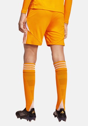 Homme portant un short de sport orange, des chaussettes montantes assorties avec des rayures blanches, et des crampons noirs, debout devant un fond uni.