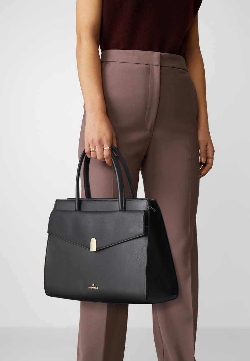 Femme tenant un sac à main en cuir noir avec fermoir doré, portant un pantalon marron et un haut foncé sur un fond uni.