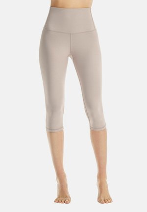 Beige Leggings mit hoher Taille und knielanger Länge an einer stehenden Person mit nackten Füßen vor einem schlichten hellen Hintergrund.