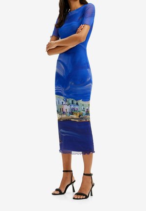 Frau trägt ein eng anliegendes blaues Midikleid mit einem Küstenstadtmotiv am Saum und schwarze, hochhackige Sandalen mit Knöchelriemen.