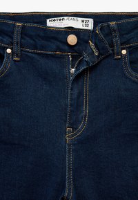 Donkerblauwe denim jeans met een rits en sluiting met messing knop. Gele stiksels langs de naden, vijf-pocket ontwerp, zonder patronen.
