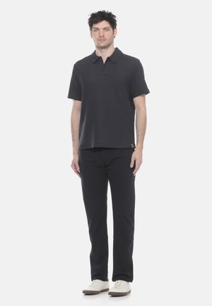 Uomo in piedi che indossa una polo nera a maniche corte, pantaloni neri e scarpe da ginnastica bianche su uno sfondo chiaro e uniforme.